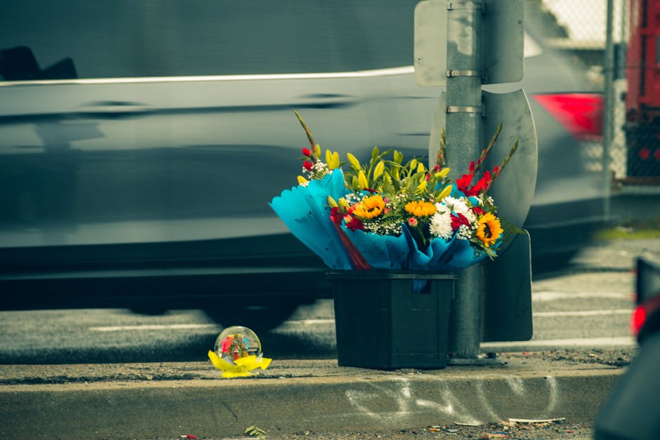 A floral arrangement of fresh flowers including sunflowers, yellow lilies, red gladioli, and white daisies, wrapped in bright blue paper, is placed in a black plastic container on the pavement beside