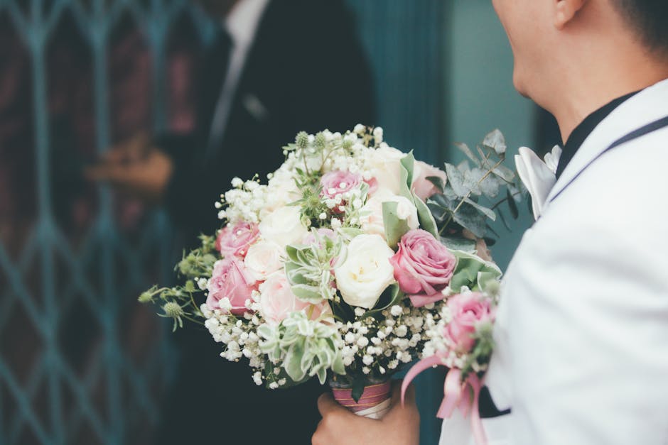 A close-up view of a floral arrangement held by a man dressed in a white suit and black bow tie, featuring a bouquet of fresh flowers including pink roses, white roses, and delicate white baby's breat