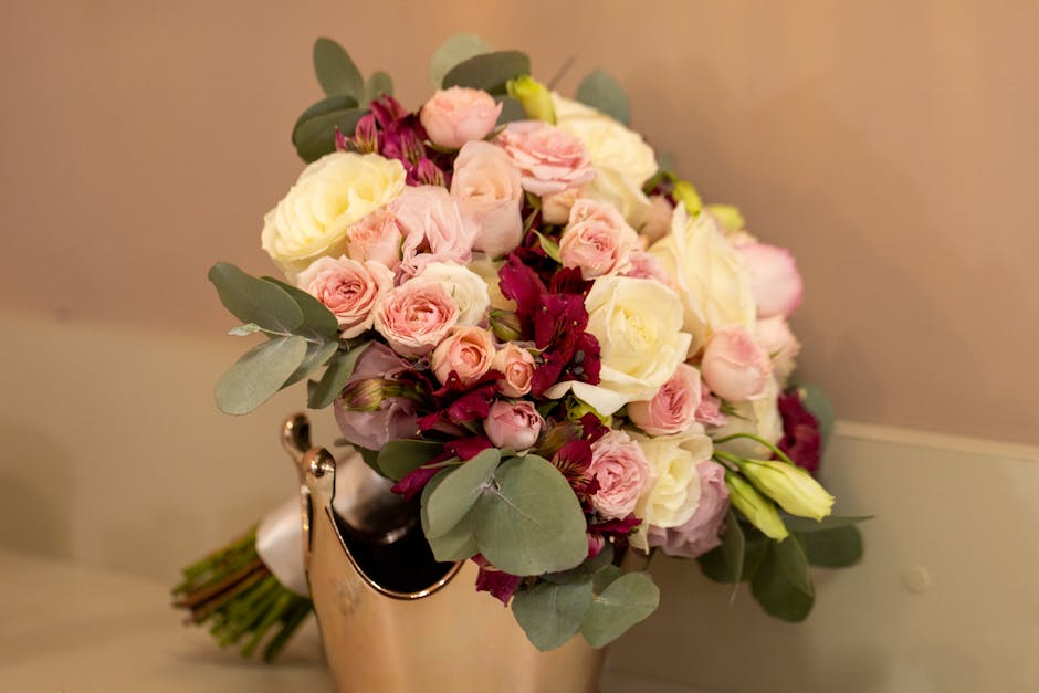 A floral arrangement featuring a bouquet of fresh roses in shades of soft pink and creamy white, interspersed with smaller pink buds and deep burgundy flowers. The bouquet is accented with broad, silv