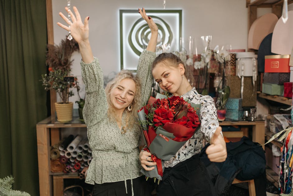 Two young women celebrating with a floral gift in a florist shop. The woman on the right is holding a bouquet of vibrant red roses, wrapped in grey and red paper, with a red ribbon, while giving a thu
