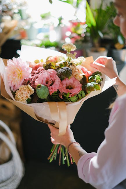 A person holding a large bouquet of fresh flowers wrapped in cream and white paper, featuring pink and white roses, peonies, and carnations with green foliage, in a well-lit indoor setting. The bouque