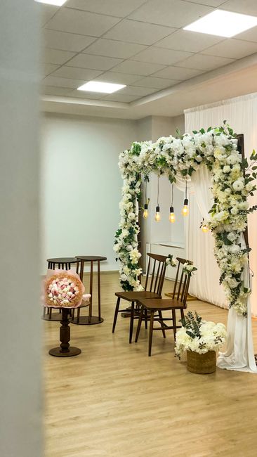 A floral arrangement featuring a large, rectangular arch draped with white fabric and richly adorned with an abundance of white roses, peonies, and other soft white blooms, complemented by greenery ac