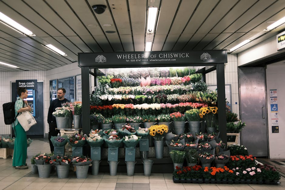 A pop-up flower stall with a black overhead sign reading 'Wheeler of Chiswick' displays a vibrant arrangement of fresh flowers, including roses, lilies, and sunflowers, organized in metal buckets and